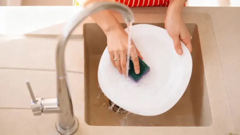 Persona che lava un piatto bianco a mano con una spugna sotto l'acqua corrente