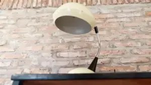 Vecchia lampada da tavolo con paralume chiaro e base curva, posizionata contro un muro in mattoni.