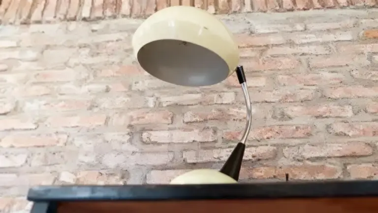 Vecchia lampada da tavolo con paralume chiaro e base curva, posizionata contro un muro in mattoni.