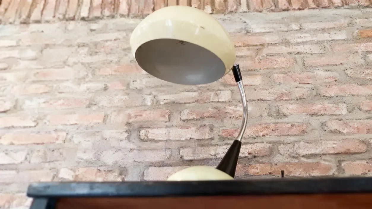 Vecchia lampada da tavolo con paralume chiaro e base curva, posizionata contro un muro in mattoni.