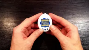 Una persona tiene in mano un Tamagotchi bianco con dettagli blu su un tavolo di legno scuro